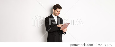 Portrait of attractive man in trendy suit, looking at digital tablet and smiling, shopping online, standing over white background Portrait of attractive man in trendy suit, looking at digital tablet and smiling, shopping online, standing over white background 98407489