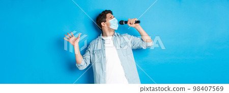 Handsome guy in medical mask singing in microphone, standing over blue background Handsome guy in medical mask singing in microphone, standing over blue background 98407569
