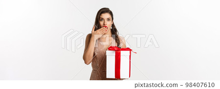 Merry Christmas. Beautiful woman looking surprised and holding gift, receiving new year presents, standing in luxury dress, standing over white background 98407610