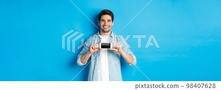 Handsome smiling man showing smartphone screen, standing against blue background for copy space Handsome smiling man showing smartphone screen, standing against blue background for copy space 98407628