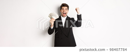 Portrait of handsome businessman in black suit, winning money and rejoicing, raising hand up with excitement, standing against white background 98407784