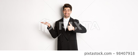 Handsome businessman in black suit, showing thumb-up and holding your product in hand over white copy space, standing against white background 98407918