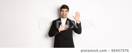 Image of handsome smiling guy telling the truth, raising on arm to promise something, standing over white background in black suit 98407976