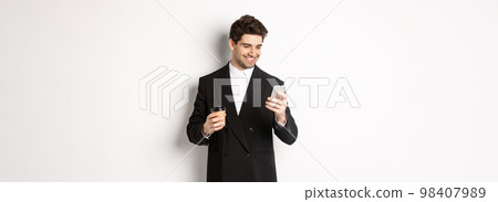 Portrait of handsome, confident businessman in black suit, drinking coffee and using mobile phone, smiling pleased, standing over white background 98407989