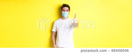 Concept of coronavirus, pandemic and social distancing. Confident young man in medical mask showing thumbs up, yellow background Concept of coronavirus, pandemic and social distancing. Confident young man in medical mask showing thumbs up, yellow background 98408046