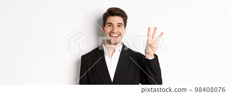 Close-up of handsome businessman in black suit, smiling amazed, showing number three, standing over white background 98408076