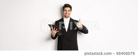Portrait of confident businessman in black suit, holding clipboard with documents and showing thumb-up in approval, praise good job, standing against white background 98408079