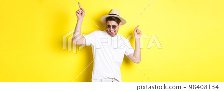 Tourism, travelling and holidays concept. Male tourist enjoying vacation, dancing in straw hat and sunglasses, pointing fingers sideways Tourism, travelling and holidays concept. Male tourist enjoying vacation, dancing in straw hat and sunglasses, pointing fingers sideways 98408134