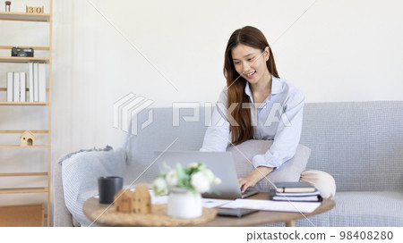Woman using laptop to work or do homework at home with smiling face in her living room, Creating happiness at work with a smile, Live performance or video call with laptop, Work from home. Woman using laptop to work or do homework at home with smiling face in her living room, Creating happiness at work with a smile, Live performance or video call with laptop, Work from home. 98408280