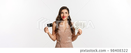 Shopping concept. Young woman deciding what to buy, looking thoughtful at upper left corner and showing credit card with money, standing in party dress Shopping concept. Young woman deciding what to buy, looking thoughtful at upper left corner and showing credit card with money, standing in party dress 98408322