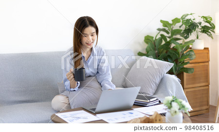 Beautiful Asian woman working on laptop and sipping coffee with smiling face in her home, Creating happiness at work with a smile, Freelancer working at home happily, Work from home. Beautiful Asian woman working on laptop and sipping coffee with smiling face in her home, Creating happiness at work with a smile, Freelancer working at home happily, Work from home. 98408516