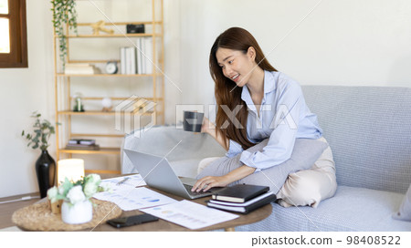 Beautiful Asian woman working on laptop and sipping coffee with smiling face in her home, Creating happiness at work with a smile, Freelancer working at home happily, Work from home. Beautiful Asian woman working on laptop and sipping coffee with smiling face in her home, Creating happiness at work with a smile, Freelancer working at home happily, Work from home. 98408522