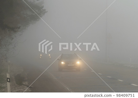 Car traveling on the foggy road with headlights or headlamps on. Low visibility - Dangerous driving of cars in winter in bad weather. 98409751