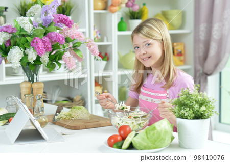 little girl preparing fresh salad on kitchen table with tablet at home 98410076