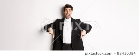 Portrait of super happy man showing christmas promo offer, pointing fingers down and smiling amazed, wearing formal party suit, standing over white background Portrait of super happy man showing christmas promo offer, pointing fingers down and smiling amazed, wearing formal party suit, standing over white background 98410384