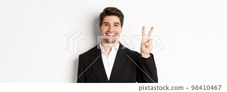 Close-up of handsome businessman in black suit, smiling amazed, showing number two, standing over white background 98410467