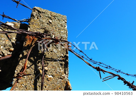 Barbed wire, double wire, metal tape with sharp spikes for barriers. Rusty barbed wire against the blue sky. The concept of war, restriction of rights and freedoms. Concrete pillar. 98410563