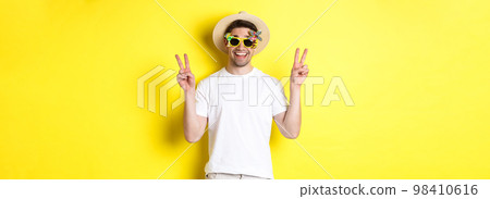 Concept of tourism and lifestyle. Happy man enjoying trip, wearing summer hat and sunglasses, posing with peace signs for photo, yellow background Concept of tourism and lifestyle. Happy man enjoying trip, wearing summer hat and sunglasses, posing with peace signs for photo, yellow background 98410616