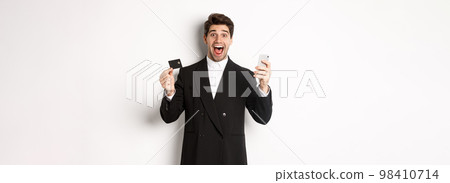 Handsome businessman in black suit smiling, showing credit card and money, looking amazed, standing against white background Handsome businessman in black suit smiling, showing credit card and money, looking amazed, standing against white background 98410714