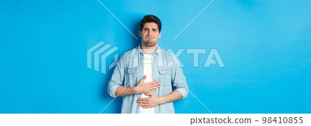 Man holding hands on belly and grimacing from pain, complaining on stomach ache, standing against blue background Man holding hands on belly and grimacing from pain, complaining on stomach ache, standing against blue background 98410855