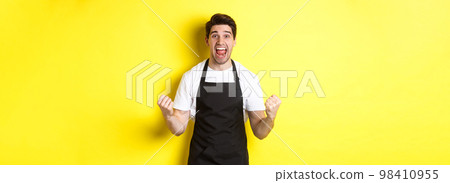 Excited coffee shop owner in black apron celebrating, making fist pump and shouting for joy, achieve goal, standing against yellow background Excited coffee shop owner in black apron celebrating, making fist pump and shouting for joy, achieve goal, standing against yellow background 98410955