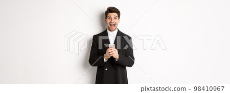 Image of excited handsome guy receiving promo offer on mobile phone, looking amazed and smiling, standing against white background Image of excited handsome guy receiving promo offer on mobile phone, looking amazed and smiling, standing against white background 98410967