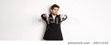 Skeptical and displeased man in black suit, looking away reluctant, showing thumbs-down, dislike something, standing over white background 98411016