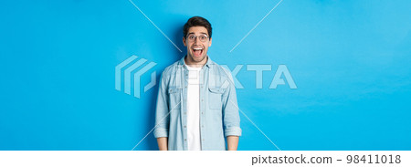Attractive happy man in glasses looking surprised, checking out advertisement, standing over blue background 98411018
