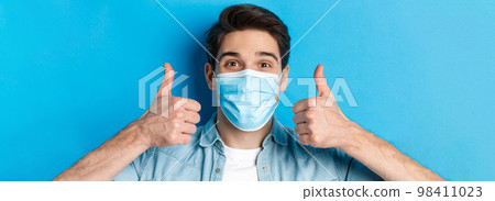 Concept of covid-19, pandemic and quarantine. Close-up of cheerful young man in medical mask smiling, showing thumbs up in approval, like and agree, standing over blue background 98411023