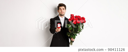 Handsome smiling man in black suit, holding roses and engagement ring, making a proposal to marry him, standing against white background 98411126