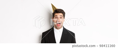 Close-up of excited handsome guy in trendy suit and party hat, looking amazed, celebrating winter holidays, standing against white background 98411160