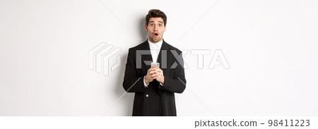 Amazed handsome guy in black suit reacting to cool promo offer, holding mobile phone, standing against white background 98411223