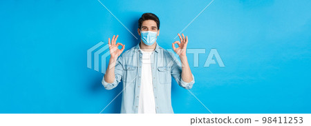 Concept of covid-19, pandemic and social distancing. Satisfied man in medical mask, showing ok signs in approval, agree or like something good, standing over blue background 98411253