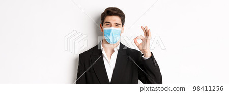 Concept of covid-19, business and social distancing. Close-up of good-looking businessman in trendy suit and medical mask, showing okay sign and winking, standing against white background 98411256