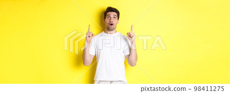 Impressed guy checking out promo offer, looking and pointing fingers up at advertisement, standing over yellow background Impressed guy checking out promo offer, looking and pointing fingers up at advertisement, standing over yellow background 98411275