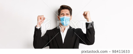 Concept of covid-19, business and social distancing. Close-up of handsome man in suit and medical mask, rejoicing and winning, raising hands up, shouting yes 98411633