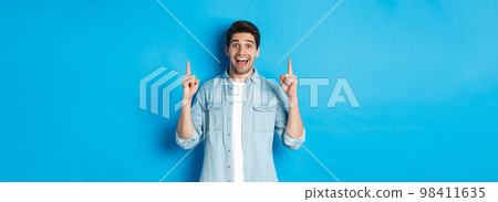 Portrait of happy 25s guy with beard, pointing fingers up and smiling, showing advertisement, standing against blue background 98411635