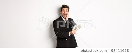 Image of rich and happy man in black suit, winning prize, holding money and looking excited at camera, standing over white background 98411638