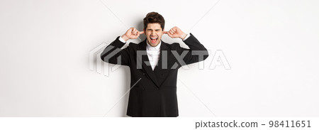 Portrait of annoyed and bothered businessman in black suit, shut ears and yelling, complaining loud noise, standing against white background Portrait of annoyed and bothered businessman in black suit, shut ears and yelling, complaining loud noise, standing against white background 98411651