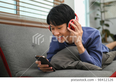 Happy young asian man listening to music in headphone and using smart phone on comfortable couch 98412126