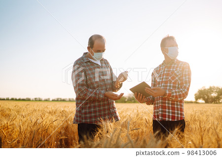 Two farmers in sterile medical masks with a tablet in their hands in a wheat field during pandemic. Two farmers in sterile medical masks with a tablet in their hands in a wheat field during pandemic. 98413006