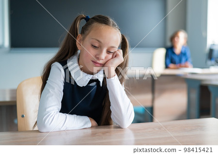 Little caucasian girl is bored at the lesson at school. The schoolgirl is sitting at her desk and the teacher is sitting in the background. Little caucasian girl is bored at the lesson at school. The schoolgirl is sitting at her desk and the teacher is sitting in the background. 98415421