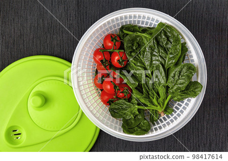 Washed red cherry tomatoes on a branch and green spinach leaves in a centrifuge strainer on a table with a black textile tablecloth 98417614