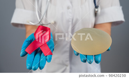 Doctor mammologist is holding a silicone breast implant and a pink ribbon. 98420230
