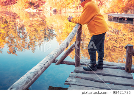 A man stands on a wooden deck leaning on a railing and looks at a forest lake A man stands on a wooden deck leaning on a railing and looks at a forest lake 98421785