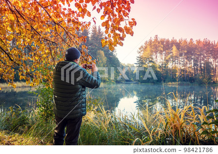 A man stands on the picturesque lakeshore in the evening. The man takes a photo of the lake with a mobile phone A man stands on the picturesque lakeshore in the evening. The man takes a photo of the lake with a mobile phone 98421786