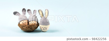 Happy Easter concept. Three Easter eggs with funny faces and gray crocheted hats with bunny ears in a decorative nest of sisal on a blue background. Banner 98422256