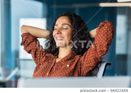Close up pjoto. A young beautiful Latin American businesswoman sits in the office at a table with a laptop, hands behind her head, eyes closed, resting, smiling. Close up pjoto. A young beautiful Latin American businesswoman sits in the office at a table with a laptop, hands behind her head, eyes closed, resting, smiling. 98422889