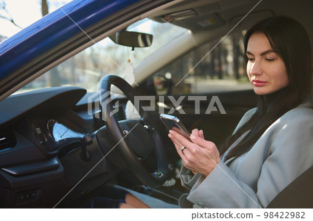 Confident successful middle-aged businesswoman, dressed in winter coat, sitting in the car on driver's seat, and using smart phone, making call to clients, planning business meetings. People Business Confident successful middle-aged businesswoman, dressed in winter coat, sitting in the car on driver's seat, and using smart phone, making call to clients, planning business meetings. People Business 98422982