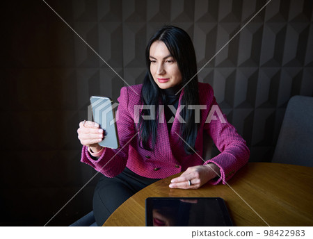 Confident middle-aged Caucasian businesswoman, dressed in elegant formal wear, using her modern smartphone, having an online business conference with her colleagues or clients, from a cafe interior Confident middle-aged Caucasian businesswoman, dressed in elegant formal wear, using her modern smartphone, having an online business conference with her colleagues or clients, from a cafe interior 98422983